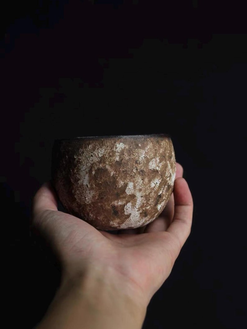 May include: A small, round, ceramic bowl with a textured, brown surface and white accents. The bowl is held in a hand against a dark background. The bowl's shape is simple and organic, suggesting a handmade quality.