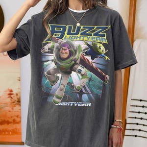 Op de afbeelding: Donkergrijs T-shirt met een afbeelding van Buzz Lightyear en een alien uit de film Lightyear. Het ontwerp bevat de tekst "BUZZ LIGHTYEAR" in geel en blauw. Het shirt heeft een relaxte, oversized pasvorm.