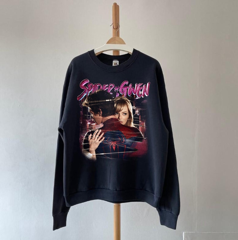 May include: A navy blue sweatshirt with a graphic of two people embracing, and the text "Spider & Gwen" in pink and white. The image has a vintage, faded appearance. The sweatshirt is hanging on a wooden hanger.