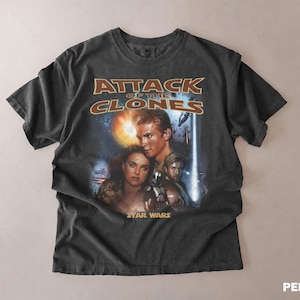 May include: Dark grey t-shirt with the movie poster for "Attack of the Clones" from the Star Wars franchise. The design features characters and the movie title in gold and orange lettering.