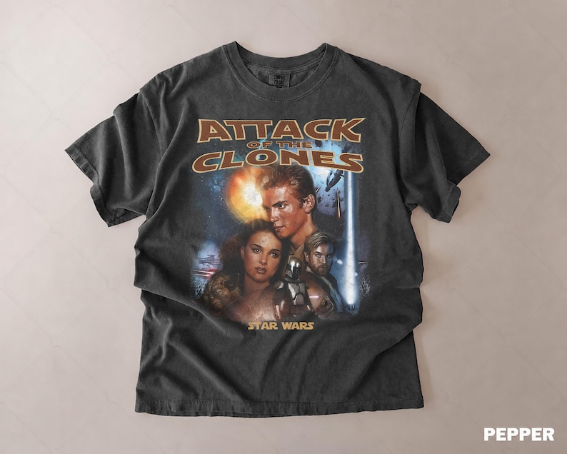 May include: Dark grey t-shirt with the movie poster for "Attack of the Clones" from the Star Wars franchise. The design features characters and the movie title in gold and orange lettering.