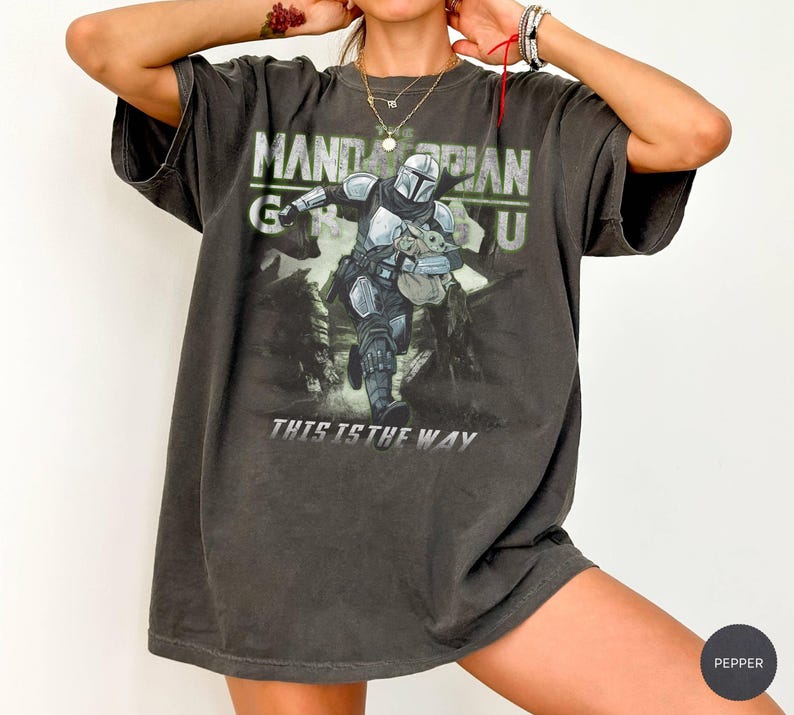 Puede incluir: Camiseta gris oscuro con un dise&ntilde;o de The Mandalorian y Grogu de Star Wars. El dise&ntilde;o incluye el texto "MANDALORIAN" y "THIS IS THE WAY" en blanco y verde. La camiseta tiene un aspecto vintage y desgastado.
