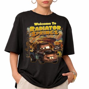 Puede incluir: Camiseta negra con un dise&ntilde;o de la pel&iacute;cula Cars. El dise&ntilde;o incluye el texto "Welcome To Radiator Springs" en amarillo y naranja, con una imagen de Mate. La camiseta es de cuello redondo y manga corta.