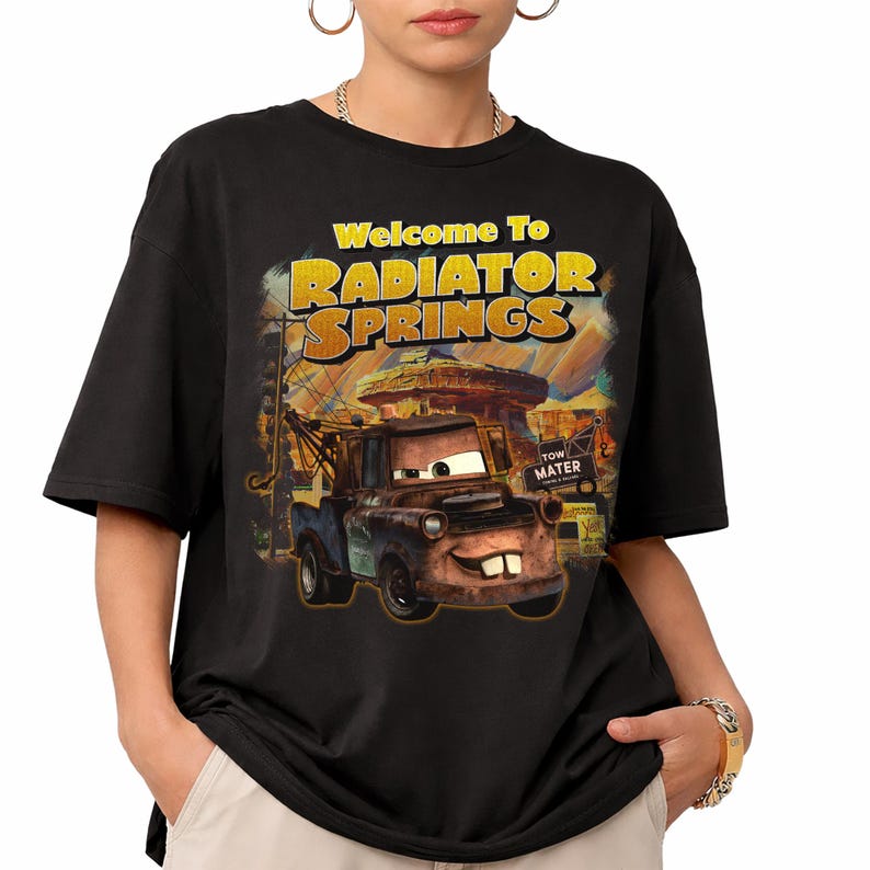 Puede incluir: Camiseta negra con un dise&ntilde;o de la pel&iacute;cula Cars. El dise&ntilde;o incluye el texto "Welcome To Radiator Springs" en amarillo y naranja, con una imagen de Mate. La camiseta es de cuello redondo y manga corta.