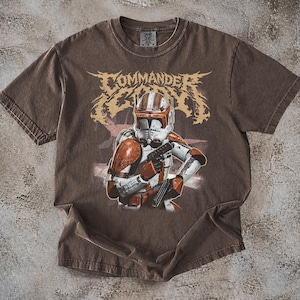 May include: Brown t-shirt featuring a graphic of a soldier in white and orange armor, holding a weapon. The word "COMMANDER" is printed above the soldier in a stylized font. The shirt has a vintage, worn look.