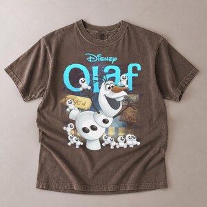 May include: Brown t-shirt featuring the Disney character Olaf from Frozen. The design includes Olaf, small snowmen, and the word "Olaf" in blue. The shirt has a vintage, washed look.