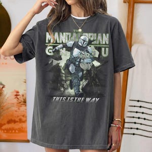 Puede incluir: Camiseta gris oscuro con un gr&aacute;fico de The Mandalorian y Grogu. El dise&ntilde;o incluye el texto "The Mandalorian" y "This is the Way" en blanco y verde. La camiseta tiene un aspecto vintage y desgastado.