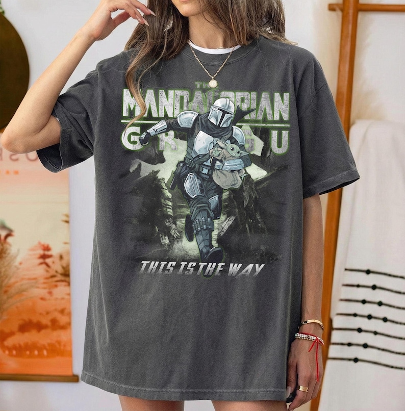 Puede incluir: Camiseta gris oscuro con un gr&aacute;fico de The Mandalorian y Grogu. El dise&ntilde;o incluye el texto "The Mandalorian" y "This is the Way" en blanco y verde. La camiseta tiene un aspecto vintage y desgastado.