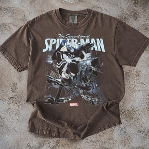 May include: Brown t-shirt featuring a graphic of Spider-Man in black and white, with the text "The Sensational Spider-Man" in white. The Marvel logo is at the bottom. The shirt is made of a soft material.