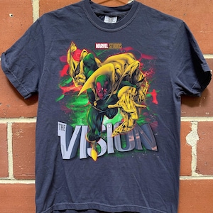 Puede incluir: Camiseta azul marino con un gr&aacute;fico del personaje Vision de Marvel Studios. El dise&ntilde;o incluye a Vision en una pose din&aacute;mica con acentos verdes, rojos y amarillos, y el texto "THE VISION".