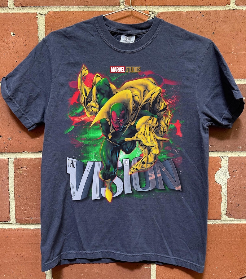 Puede incluir: Camiseta azul marino con un gr&aacute;fico del personaje Vision de Marvel Studios. El dise&ntilde;o incluye a Vision en una pose din&aacute;mica con acentos verdes, rojos y amarillos, y el texto "THE VISION".