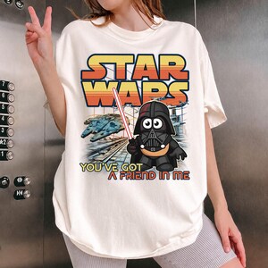 Może przedstawiać: Kremowy t-shirt z nadrukiem graficznym zawierającym tekst STAR WARS oraz YOU'VE GOT A FRIEND IN ME. Wzór przedstawia postać Dartha Vadera w stylu kreskówkowym z czerwonym mieczem świetlnym, statek kosmiczny oraz stację kosmiczną w tle.