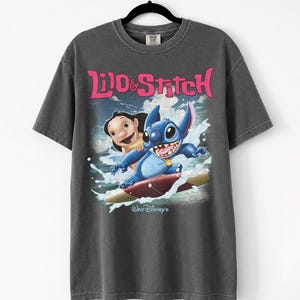 Peut inclure: T-shirt gris fonc&eacute; avec l'inscription rose "Lilo & Stitch" et une image de Lilo et Stitch surfant. Le logo Walt Disney est en bas. Le t-shirt est suspendu &agrave; un cintre noir.