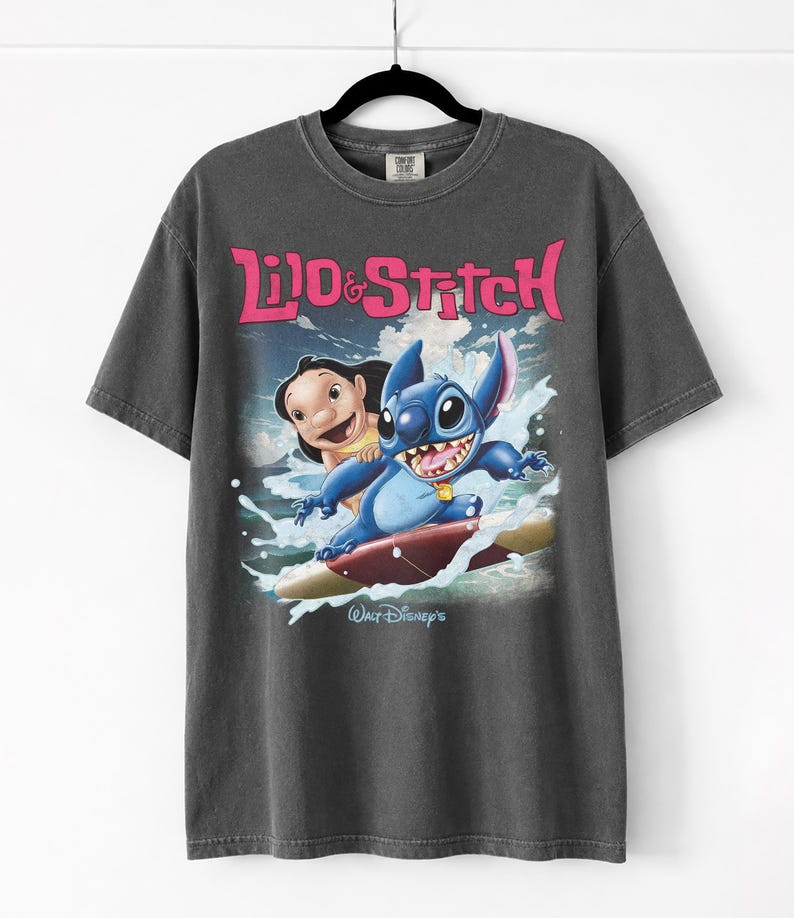 Peut inclure: T-shirt gris fonc&eacute; avec l'inscription rose "Lilo & Stitch" et une image de Lilo et Stitch surfant. Le logo Walt Disney est en bas. Le t-shirt est suspendu &agrave; un cintre noir.