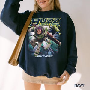 Op de afbeelding: Marineblauwe sweatshirt met een afbeelding van Buzz Lightyear en een alien uit de film Lightyear. De sweatshirt toont de woorden "BUZZ LIGHTYEAR" in geel en groen. Het woord "LIGHTYEAR" staat ook onderaan de afbeelding.