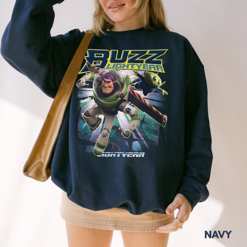 Op de afbeelding: Marineblauwe sweatshirt met een afbeelding van Buzz Lightyear en een alien uit de film Lightyear. De sweatshirt toont de woorden "BUZZ LIGHTYEAR" in geel en groen. Het woord "LIGHTYEAR" staat ook onderaan de afbeelding.