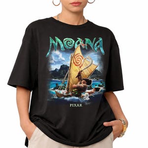 Pu&ograve; includere: T-shirt nera con grafica del film "Moana". Il design include il titolo del film in turchese, una barca a vela con un personaggio e la scritta "PIXAR" in basso. La maglietta ha maniche corte e girocollo.