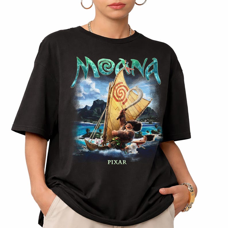 Pu&ograve; includere: T-shirt nera con grafica del film "Moana". Il design include il titolo del film in turchese, una barca a vela con un personaggio e la scritta "PIXAR" in basso. La maglietta ha maniche corte e girocollo.