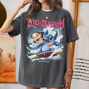 Peut inclure: T-shirt gris fonc&eacute; avec les personnages Lilo et Stitch surfant. Le graphisme comprend le texte "Lilo & Stitch" en rose et "Walt Disney's" en blanc. Le t-shirt est de coupe ample.