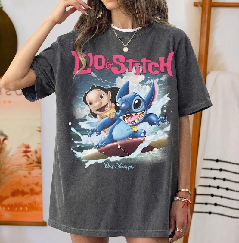 Peut inclure: T-shirt gris fonc&eacute; avec les personnages Lilo et Stitch surfant. Le graphisme comprend le texte "Lilo & Stitch" en rose et "Walt Disney's" en blanc. Le t-shirt est de coupe ample.