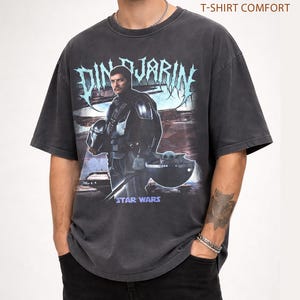 May include: Dark gray t-shirt featuring a graphic of Din Djarin from Star Wars, with the text "DIN DJARIN" in a stylized font. The shirt also includes the text "STAR WARS" and "PEPPER T-SHIRT COMFORT".