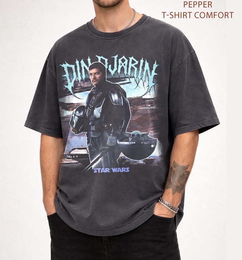 May include: Dark gray t-shirt featuring a graphic of Din Djarin from Star Wars, with the text "DIN DJARIN" in a stylized font. The shirt also includes the text "STAR WARS" and "PEPPER T-SHIRT COMFORT".