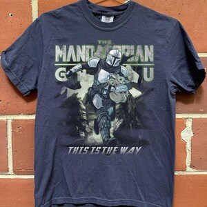 Puede incluir: Camiseta azul marino con un gr&aacute;fico de The Mandalorian y Grogu. El dise&ntilde;o incluye el texto "THE MANDALORIAN GROGU" y "THIS IS THE WAY". La imagen muestra al Mandaloriano corriendo con Grogu en una bolsa.