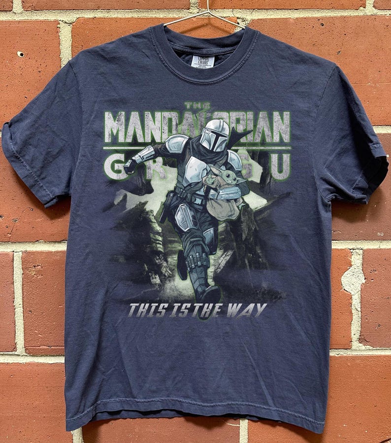 Puede incluir: Camiseta azul marino con un gr&aacute;fico de The Mandalorian y Grogu. El dise&ntilde;o incluye el texto "THE MANDALORIAN GROGU" y "THIS IS THE WAY". La imagen muestra al Mandaloriano corriendo con Grogu en una bolsa.