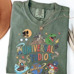 Może przedstawiać: Oliwkowozielony t-shirt z nadrukiem graficznym z napisem Universal Studios. Wzór zawiera sylwetkę E.T. na rowerze, zamek Hogwart, tyranozaura, Minionka, Optimusa Prime'a, taśmę filmową oraz logo kuli ziemskiej wytwórni Universal.