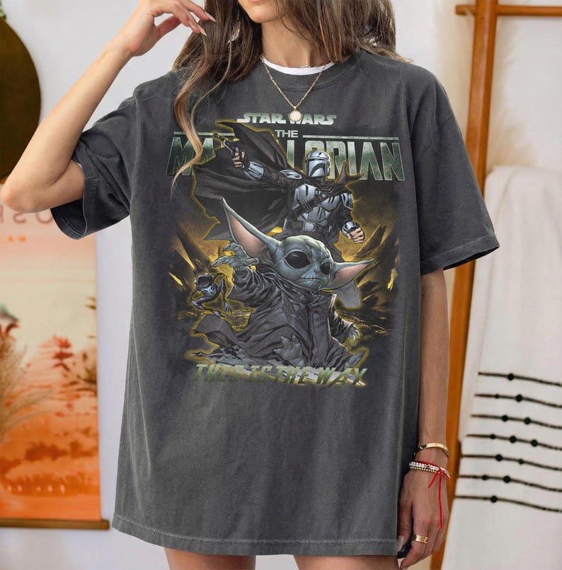 May include: Dark gray t-shirt featuring a graphic from the Star Wars series, "The Mandalorian." The design includes the Mandalorian, Grogu, and the text "This is the Way." The shirt has a crew neck and short sleeves.