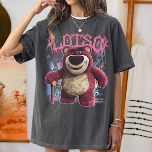 K&ouml;nnte beinhalten: Dunkelgraues T-Shirt mit einer Grafik von Lotso, einem rosa B&auml;rencharakter, und dem Wort "LOTSO" in rosa, stilisiertem Text. Der B&auml;r h&auml;lt einen Holzstab. Das Shirt hat einen Vintage-Look.