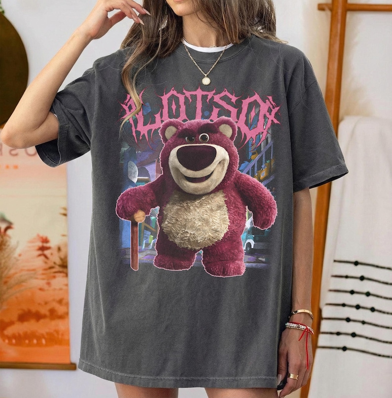 K&ouml;nnte beinhalten: Dunkelgraues T-Shirt mit einer Grafik von Lotso, einem rosa B&auml;rencharakter, und dem Wort "LOTSO" in rosa, stilisiertem Text. Der B&auml;r h&auml;lt einen Holzstab. Das Shirt hat einen Vintage-Look.