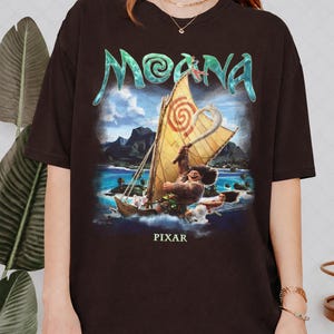 Pu&ograve; includere: Una maglietta marrone scuro con una grafica colorata del film d'animazione "Moana". Il design include il titolo del film in lettere turchesi, una barca a vela con un personaggio e la parola "PIXAR" in basso.