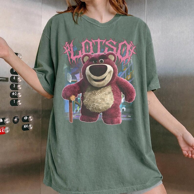 K&ouml;nnte beinhalten: Gr&uuml;nes T-Shirt mit einem Aufdruck von Lotso, einem rosa B&auml;ren, und dem Wort "LOTSO" in Rosa dar&uuml;ber. Der B&auml;r h&auml;lt einen Holzstock. Das T-Shirt hat kurze &Auml;rmel und eine lockere Passform.