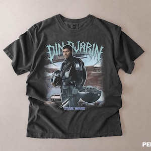 May include: Dark gray t-shirt featuring a graphic of Din Djarin from Star Wars, holding his helmet, with Grogu in a pod. The text "DIN DJARIN" is above the image, and "STAR WARS" is below. The shirt has a vintage, worn look.