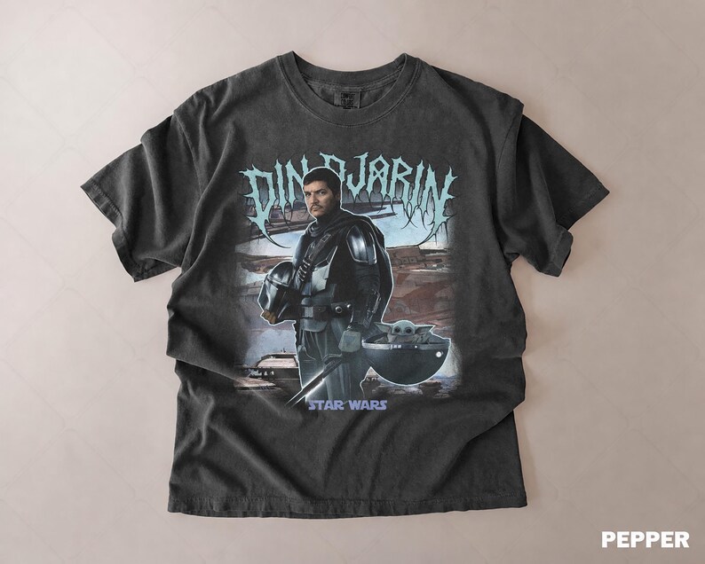 May include: Dark gray t-shirt featuring a graphic of Din Djarin from Star Wars, holding his helmet, with Grogu in a pod. The text "DIN DJARIN" is above the image, and "STAR WARS" is below. The shirt has a vintage, worn look.
