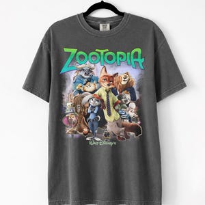 Puede incluir: Camiseta gris oscuro con la palabra "Zootopia" en verde y azul, con un gr&aacute;fico de personajes de la pel&iacute;cula de Disney. La camiseta est&aacute; colgada en una percha negra.