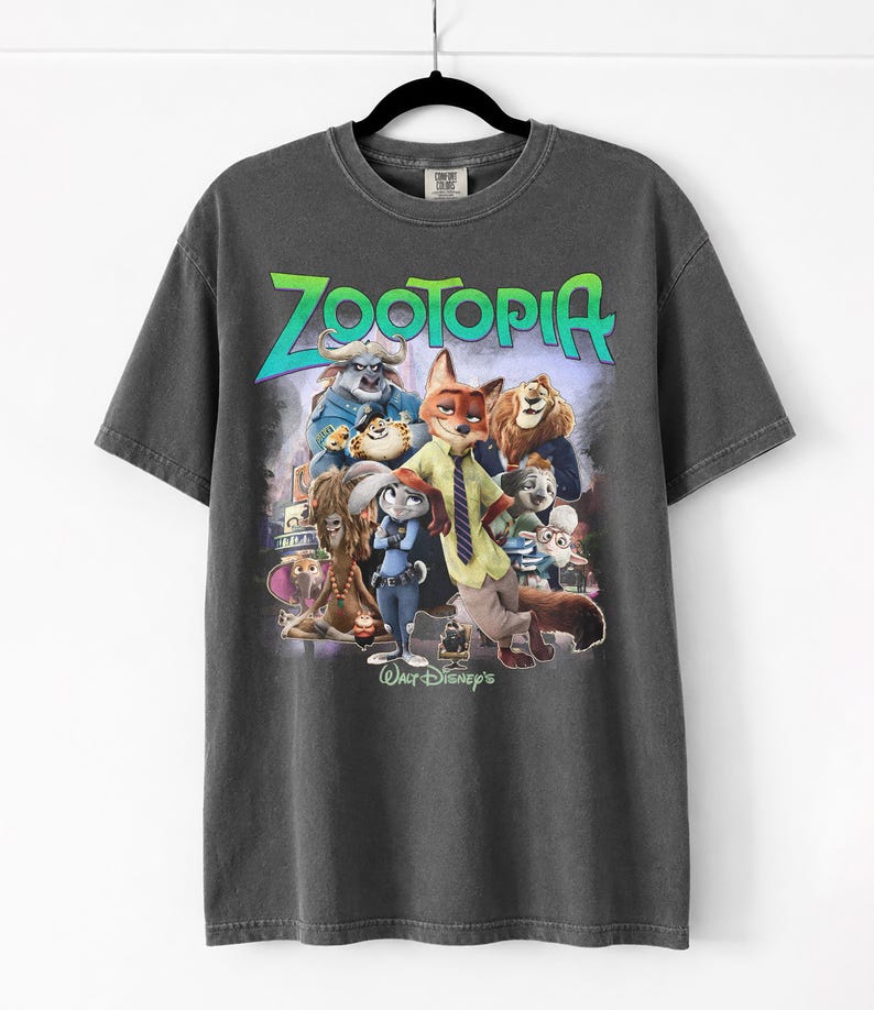 Puede incluir: Camiseta gris oscuro con la palabra "Zootopia" en verde y azul, con un gr&aacute;fico de personajes de la pel&iacute;cula de Disney. La camiseta est&aacute; colgada en una percha negra.