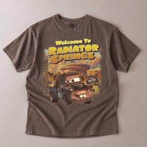 Puede incluir: Camiseta marr&oacute;n con un dise&ntilde;o de Mate de la pel&iacute;cula Cars, con el texto "Welcome To Radiator Springs". El dise&ntilde;o incluye una imagen de dibujos animados de Mate y el paisaje de Radiator Springs.