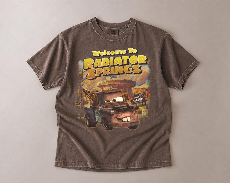 Puede incluir: Camiseta marr&oacute;n con un dise&ntilde;o de Mate de la pel&iacute;cula Cars, con el texto "Welcome To Radiator Springs". El dise&ntilde;o incluye una imagen de dibujos animados de Mate y el paisaje de Radiator Springs.