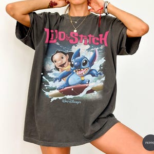 Peut inclure: T-shirt gris fonc&eacute; avec les personnages Lilo et Stitch. Le graphisme montre Lilo et Stitch surfant sur une vague, avec les mots "Lilo & Stitch" en rose. Le logo Walt Disney est en bas.