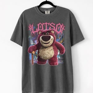 K&ouml;nnte beinhalten: Dunkelgraues T-Shirt mit einer Grafik von Lotso, einem rosa B&auml;rencharakter, und dem Wort "LOTSO" in rosa, stilisiertem Text. Der B&auml;r h&auml;lt einen Holzstock. Das T-Shirt h&auml;ngt an einem schwarzen Kleiderb&uuml;gel.