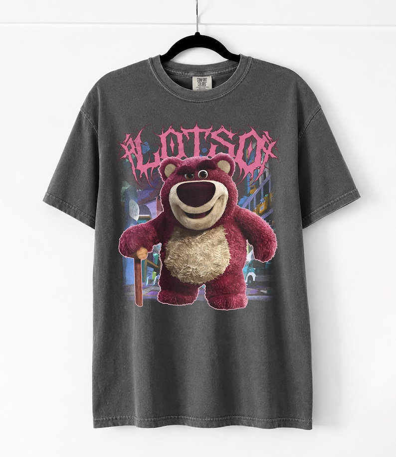 K&ouml;nnte beinhalten: Dunkelgraues T-Shirt mit einer Grafik von Lotso, einem rosa B&auml;rencharakter, und dem Wort "LOTSO" in rosa, stilisiertem Text. Der B&auml;r h&auml;lt einen Holzstock. Das T-Shirt h&auml;ngt an einem schwarzen Kleiderb&uuml;gel.