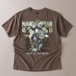 Puede incluir: Una camiseta marr&oacute;n con un gr&aacute;fico de The Mandalorian sosteniendo a Grogu, con el texto "THE MANDALORIAN GU" arriba y "THIS IS THE WAY" abajo. El dise&ntilde;o es en tonos de gris, verde y blanco.