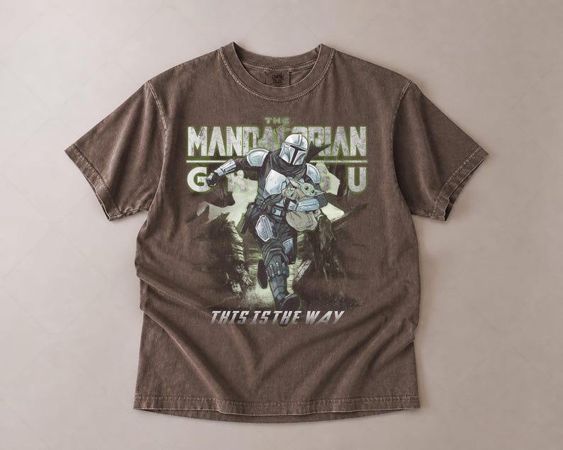 Puede incluir: Una camiseta marr&oacute;n con un gr&aacute;fico de The Mandalorian sosteniendo a Grogu, con el texto "THE MANDALORIAN GU" arriba y "THIS IS THE WAY" abajo. El dise&ntilde;o es en tonos de gris, verde y blanco.
