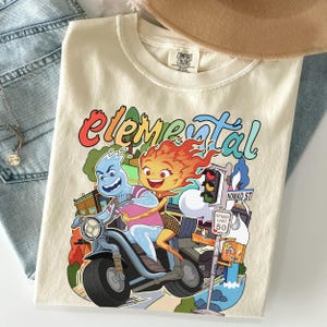Puede incluir: Una camiseta de color crema con un estampado gráfico de dos personajes de dibujos animados montando en un scooter. El texto "elemental" está impreso en una fuente colorida y estilizada sobre los personajes, junto a un semáforo y una señal.