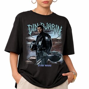 May include: Black t-shirt featuring a graphic of Din Djarin from Star Wars, holding his helmet, with Grogu in a floating pod. The text "Din Djarin" is above the image in a stylized font, with "Star Wars" below.