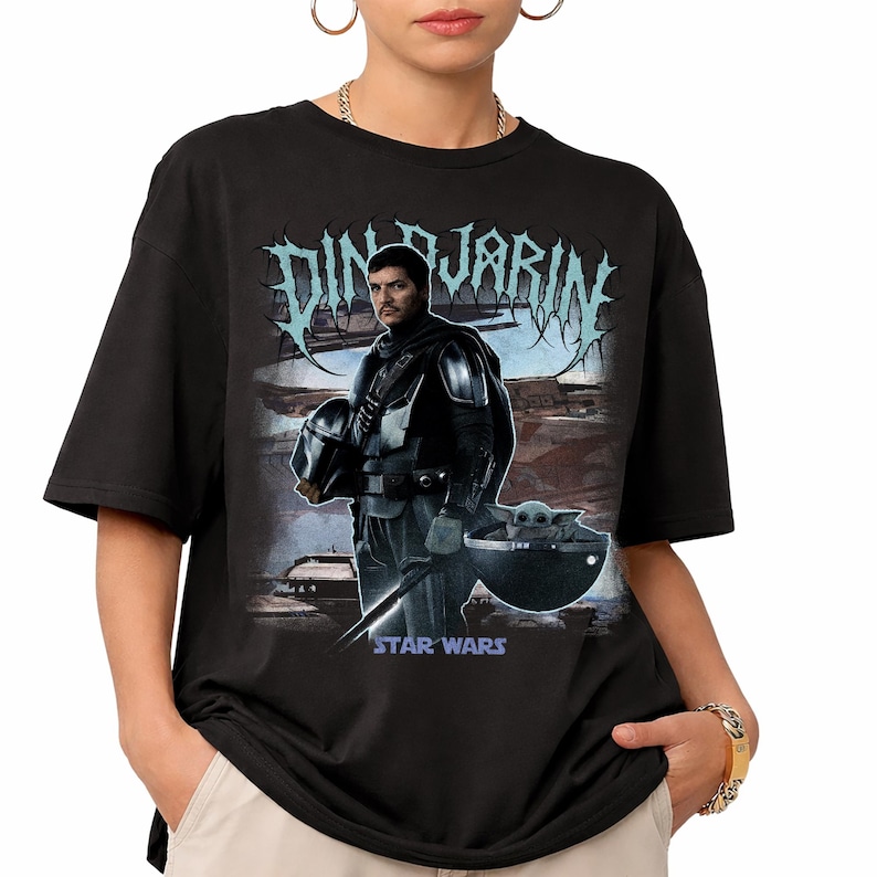 May include: Black t-shirt featuring a graphic of Din Djarin from Star Wars, holding his helmet, with Grogu in a floating pod. The text "Din Djarin" is above the image in a stylized font, with "Star Wars" below.