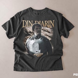 May include: Dark gray t-shirt featuring a portrait of Din Djarin from The Mandalorian. The shirt has the text "DIN DJARIN" in gold lettering above the image and "MANDALORIAN GROGU" below. The shirt has a vintage, distressed look.