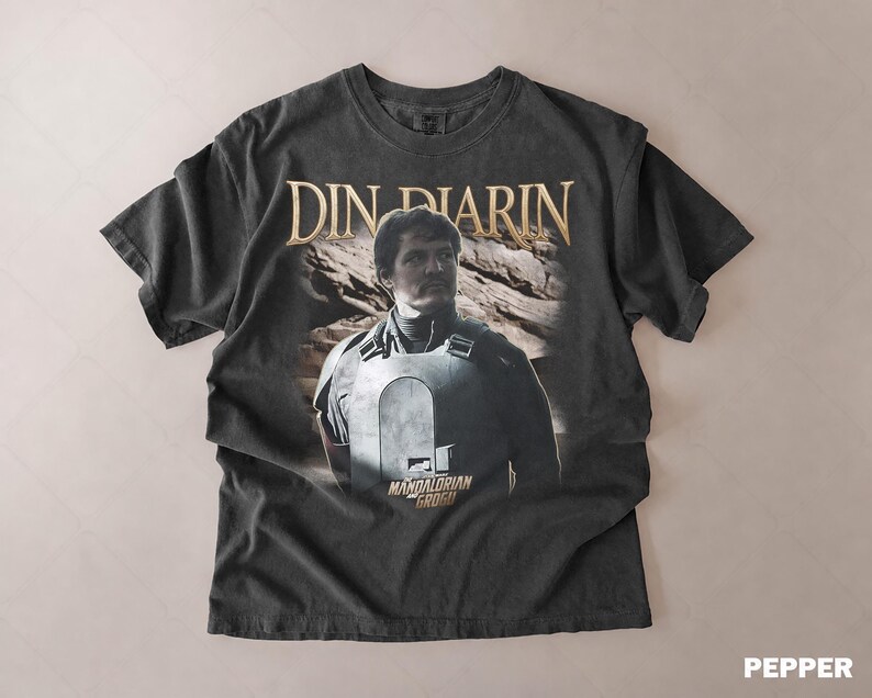 May include: Dark gray t-shirt featuring a portrait of Din Djarin from The Mandalorian. The shirt has the text "DIN DJARIN" in gold lettering above the image and "MANDALORIAN GROGU" below. The shirt has a vintage, distressed look.